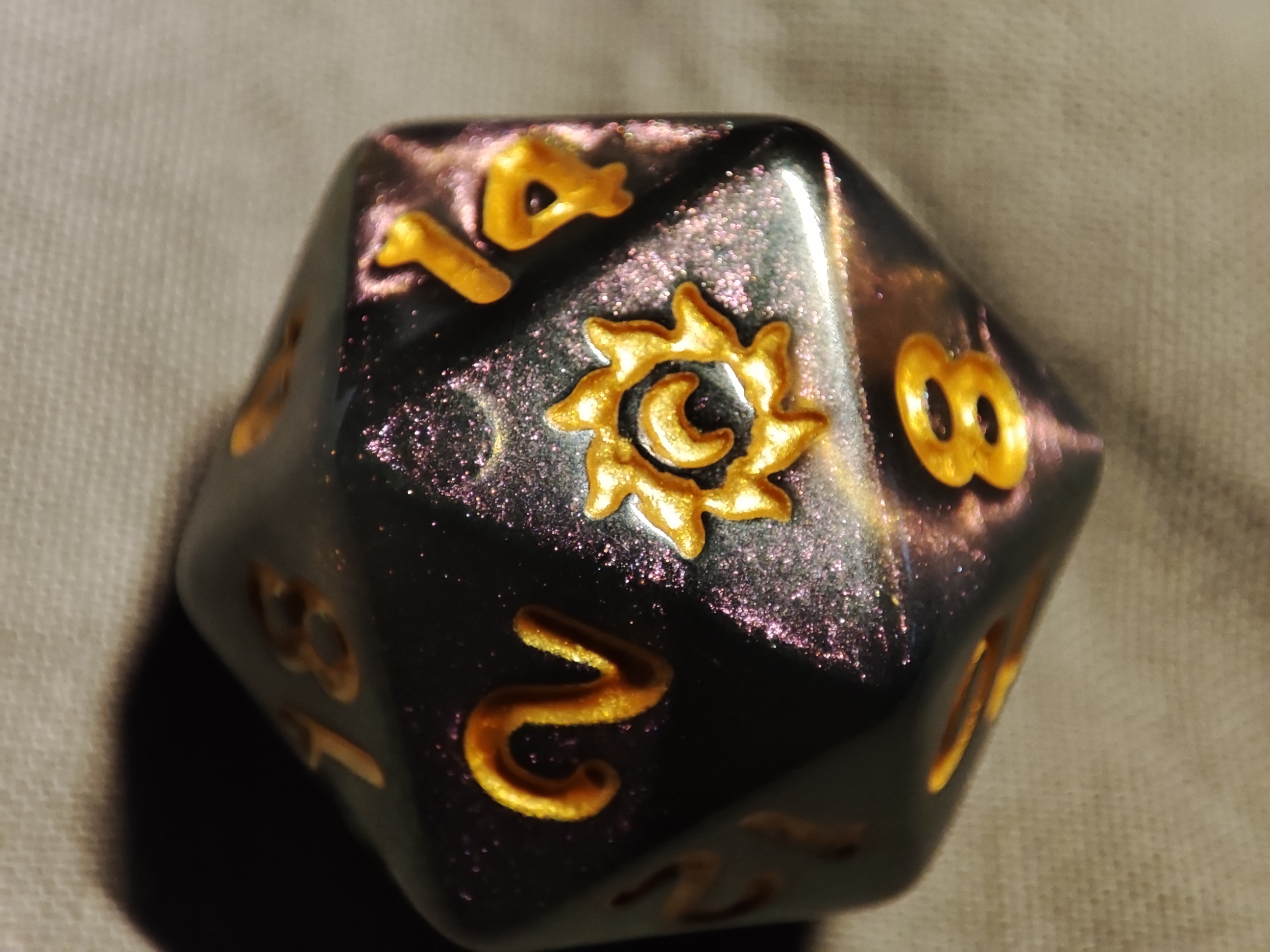 a 20-sided die, dark grey with pink glitter and gold numbers. one of the faces has a sun-moon emblem instead 