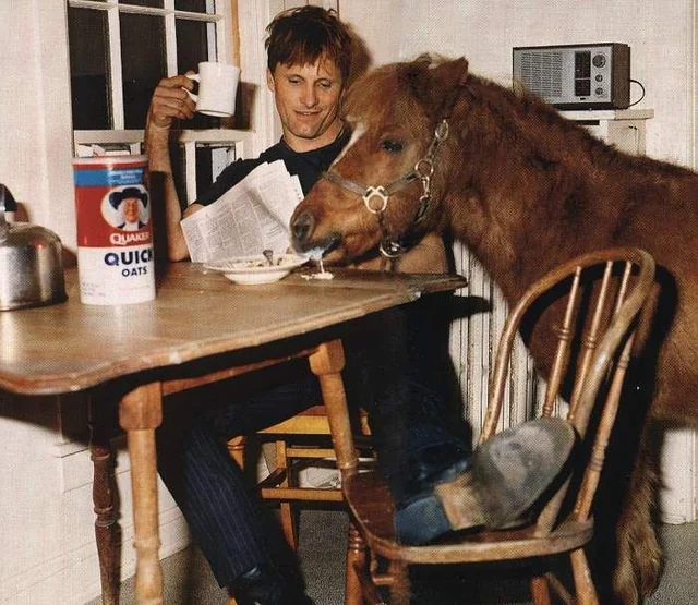 A photo of Viggo Mortensen and his horse at the breakfast table. Mortensen is holding a cup of coffee and a newspaper and is watching amusedly as his horse drinks milk out of his cereal bowl.